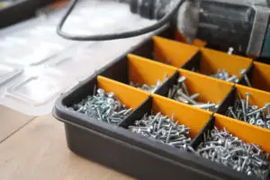 Caixa organizadora com divisórias laranja, cheia de parafusos de diferentes tamanhos e modelos. Ao fundo, uma tampa transparente e uma furadeira, sugerindo um ambiente de trabalho ou montagem. A imagem transmite organização e eficiência na escolha dos fixadores certos para cada necessidade.
