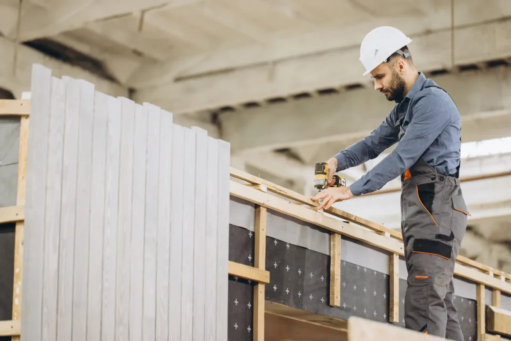 Profissional da construção civil, com capacete e parafusadeira, fixando estrutura de madeira com parafuso francês em obra modular dentro de galpão industrial.