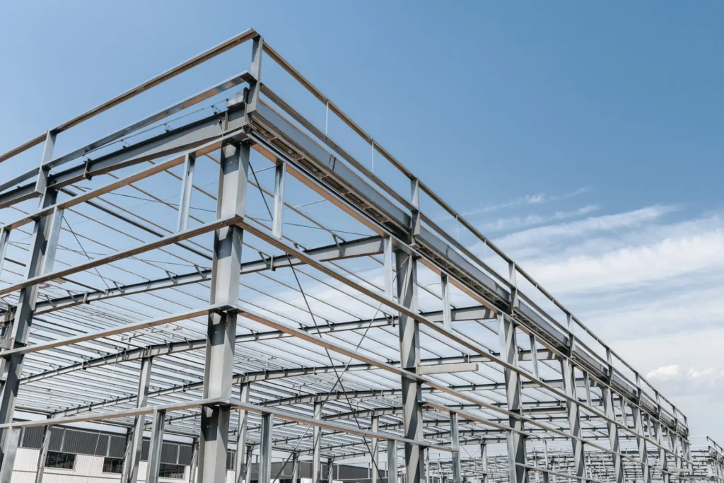 Estrutura metálica de grande porte em construção, composta por vigas e colunas de aço interligadas por parafusos de fixação, sob um céu azul claro.
